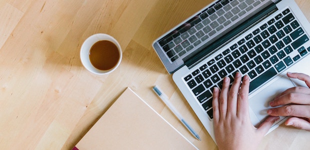 hands on a keyboard with cup of tea, pen, and journal on a table