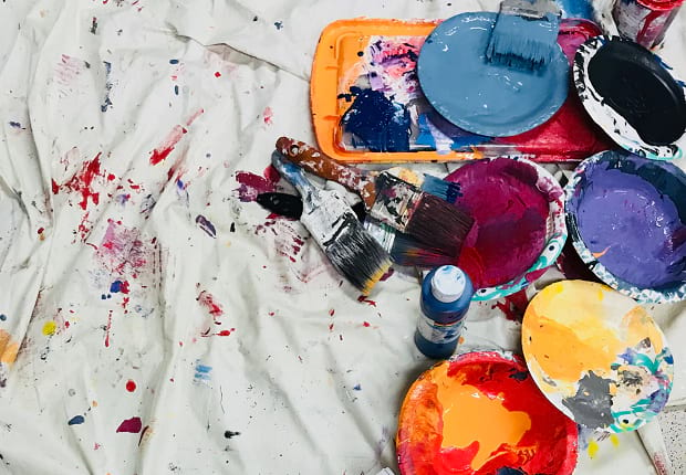 Several brushes and different colors of paint laid out on a white, paint-splattered cloth